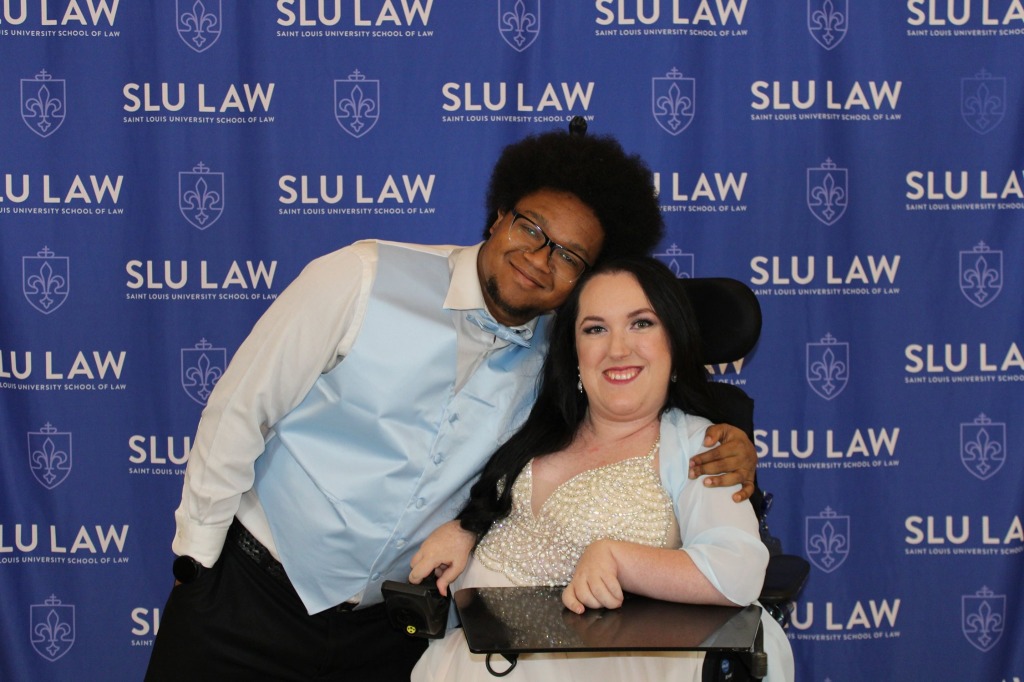 A Black man with his arm around white woman in front of a blue backdrop that says SLU LAW in white lettering. The man is wearing a white dress shirt, black dress pants, and a light blue vest and bow tie, and he wears glasses and has a goatee and afro. The woman sits in a wheelchair with a tray in front and is wearing a pale pink dress with a pearl bodice and a light blue shawl. She has long black hair.