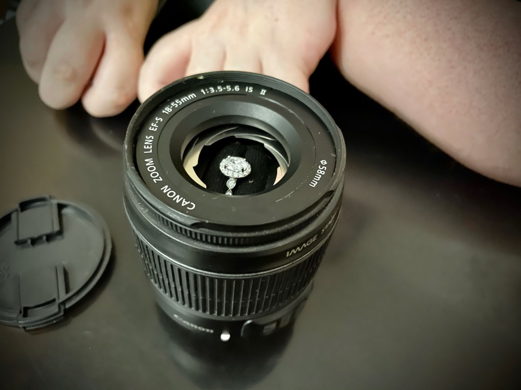 An oval-shaped diamond engagement ring with a halo of stones and a silver band inside of a customized ring box made from a Canon camera lens. White female arms are out of focus in the background and the lens cover is next to the lens box.